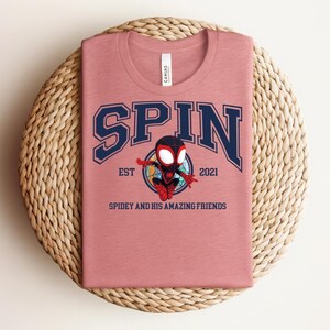 K&ouml;nnte beinhalten: Staubrosafarbenes T-Shirt mit Rundhalsausschnitt, mit dem Wort "SPIN" in marineblauen Blockbuchstaben mit wei&szlig;er Umrandung. Darunter befindet sich ein Cartoon-Bild eines Superhelden. Der Text "EST 2021" und "SPIDEY AND HIS AMAZING FRIENDS" sind ebenfalls aufgedruckt.