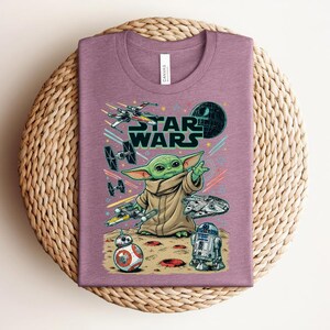Puede incluir: Una camiseta malva de cuello redondo con un estampado gr&aacute;fico de Grogu, el logotipo de Star Wars y varias naves espaciales, incluyendo un X-wing, cazas TIE, el Halc&oacute;n Milenario, BB-8 y R2-D2 sobre una superficie rocosa con estelas de luz.