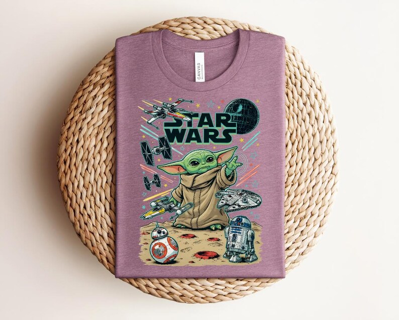 Puede incluir: Una camiseta malva de cuello redondo con un estampado gr&aacute;fico de Grogu, el logotipo de Star Wars y varias naves espaciales, incluyendo un X-wing, cazas TIE, el Halc&oacute;n Milenario, BB-8 y R2-D2 sobre una superficie rocosa con estelas de luz.