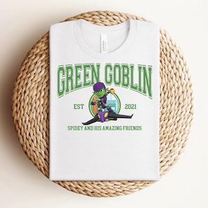 May include: White t-shirt featuring a graphic of the Green Goblin character from Spiderman. The design includes the text "GREEN GOBLIN" in green, the year "2021", and the phrase "SPIDEY AND HIS AMAZING FRIENDS". The shirt is displayed on a woven surface.