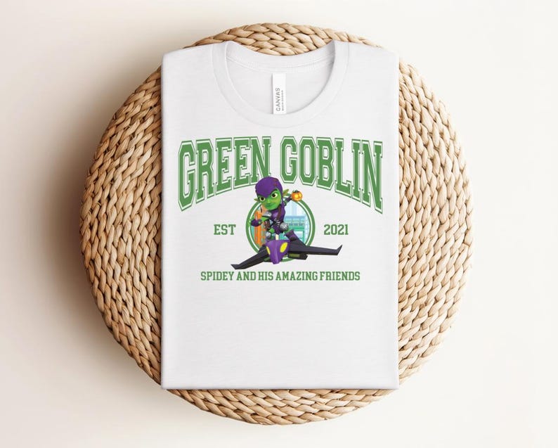 May include: White t-shirt featuring a graphic of the Green Goblin character from Spiderman. The design includes the text "GREEN GOBLIN" in green, the year "2021", and the phrase "SPIDEY AND HIS AMAZING FRIENDS". The shirt is displayed on a woven surface.