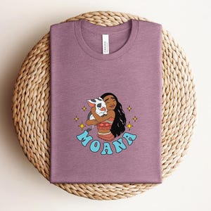 Puede incluir: Una camiseta de cuello redondo en color malva jaspeado con una ilustraci&oacute;n gr&aacute;fica de un personaje de pelo largo y oscuro abrazando a un peque&ntilde;o cerdo manchado. La palabra MOANA est&aacute; impresa en letras azul claro bajo el personaje.