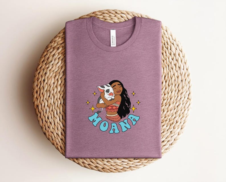Puede incluir: Una camiseta de cuello redondo en color malva jaspeado con una ilustraci&oacute;n gr&aacute;fica de un personaje de pelo largo y oscuro abrazando a un peque&ntilde;o cerdo manchado. La palabra MOANA est&aacute; impresa en letras azul claro bajo el personaje.