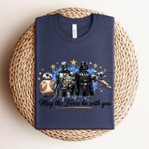 Pode incluir: Uma t-shirt azul-marinho de gola redonda com uma estampa gr&aacute;fica de BB-8, o Mandaloriano, Grogu e Darth Vader ao lado de um ca&ccedil;a estelar X-wing. O design inclui estrelas amarelas, salpicos de tinta azul e o texto May the Force be with you.