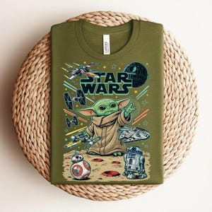 Puede incluir: Una camiseta de color verde oliva con un estampado gr&aacute;fico de Grogu de Star Wars, rodeado de varias naves espaciales, la Estrella de la Muerte, BB-8 y R2-D2. El texto STAR WARS aparece impreso en letras grandes sobre el personaje en el frente.