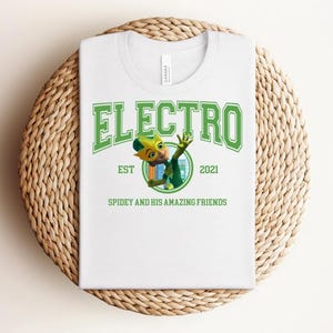 May include: White t-shirt with a green graphic that says "ELECTRO" above an illustration of a cartoon character. The text "EST 2021" and "SPIDEY AND HIS AMAZING FRIENDS" are also printed on the shirt. The shirt is folded on a woven, circular mat.
