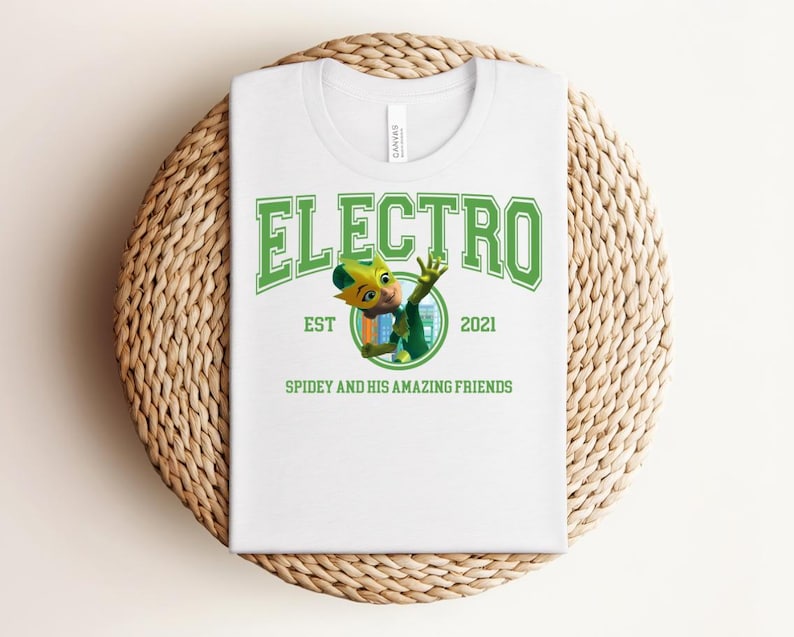 May include: White t-shirt with a green graphic that says "ELECTRO" above an illustration of a cartoon character. The text "EST 2021" and "SPIDEY AND HIS AMAZING FRIENDS" are also printed on the shirt. The shirt is folded on a woven, circular mat.