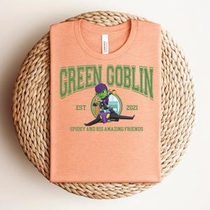 May include: Peach-colored t-shirt featuring the text "GREEN GOBLIN" in green lettering, with a cartoon illustration of the Green Goblin character. The shirt also includes the text "EST 2021" and "SPIDEY AND HIS AMAZING FRIENDS".
