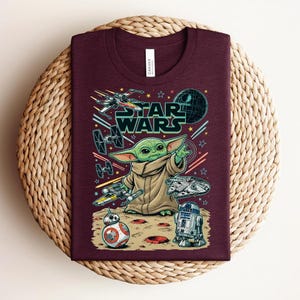 Puede incluir: Una camiseta de cuello redondo color granate con un estampado gr&aacute;fico de Star Wars. El dise&ntilde;o incluye a Grogu, BB-8, R2-D2, el Halc&oacute;n Milenario, un X-wing y un TIE fighter sobre un fondo espacial con el texto Star Wars en letras verdes.