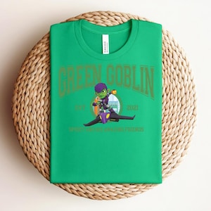 May include: A bright green t-shirt featuring the words "GREEN GOBLIN" in gold lettering. Below the text is a cartoon image of the Green Goblin riding a glider. The text "EST. 2021" and "SPIDER-MAN AND AMAZING FRIENDS" are also visible.