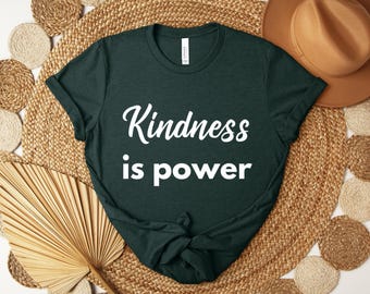 May include: Dark green t-shirt with the words "Kindness is power" in white script. The shirt is displayed on a woven mat with a brown hat and decorative fan in the background.