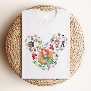K&ouml;nnte beinhalten: Wei&szlig;es T-Shirt mit einem farbenfrohen Aufdruck von Disney's Arielle, die Meerjungfrau-Figuren, angeordnet in Form eines Mickey Mouse-Kopfes. Das Design umfasst Arielle, Prinz Eric und verschiedene Meeresbewohner.