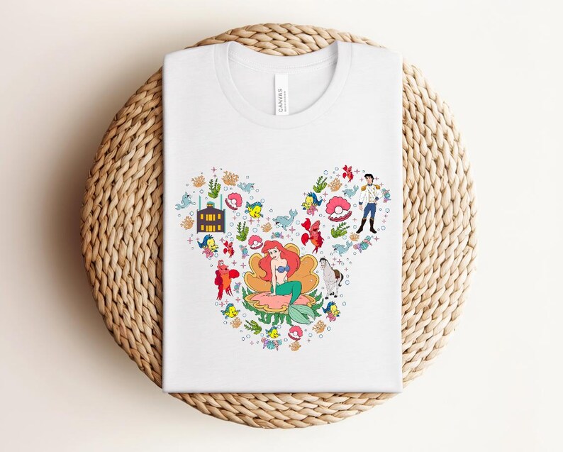 K&ouml;nnte beinhalten: Wei&szlig;es T-Shirt mit einem farbenfrohen Aufdruck von Disney's Arielle, die Meerjungfrau-Figuren, angeordnet in Form eines Mickey Mouse-Kopfes. Das Design umfasst Arielle, Prinz Eric und verschiedene Meeresbewohner.