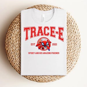 May include: White t-shirt with a red and white graphic that reads "TRACE-E" above a cartoon spider character. The text "EST 2021" and "SPIDEY AND HIS AMAZING FRIENDS" are also printed on the shirt. The shirt is displayed on a woven, circular surface.