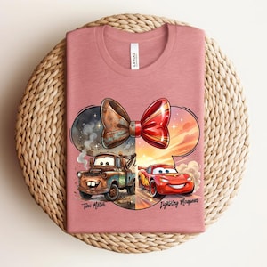 May include: A dusty rose crew neck t-shirt featuring a graphic of a mouse ear silhouette containing a rusty tow truck on the left and a red race car on the right, topped with a bow. The text reads Tow Mater and Lightning McQueen.