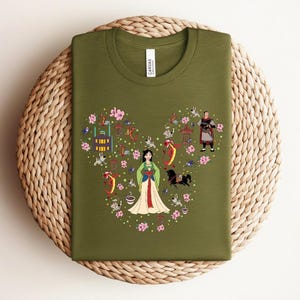 May include: Olive green t-shirt featuring a heart-shaped design filled with illustrations of a woman in a white dress, flowers, buildings, and characters. The shirt has a crew neck and a tag that says "Canvas".