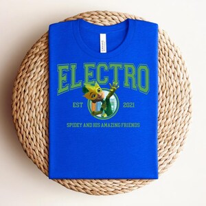 May include: A royal blue t-shirt with the word "ELECTRO" in green block letters. Below the word is a cartoon character and the text "EST 2021" and "SPIDEY AND HIS AMAZING FRIENDS". The shirt is folded on a woven mat.