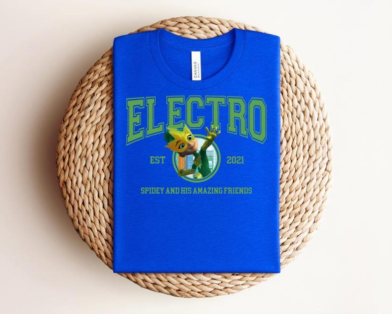 May include: A royal blue t-shirt with the word "ELECTRO" in green block letters. Below the word is a cartoon character and the text "EST 2021" and "SPIDEY AND HIS AMAZING FRIENDS". The shirt is folded on a woven mat.