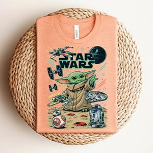 Puede incluir: Una camiseta de color melocot&oacute;n con un estampado gr&aacute;fico de Star Wars. El dise&ntilde;o incluye a Grogu, un X-wing, un caza TIE, el Halc&oacute;n Milenario, BB-8, R2-D2 y la Estrella de la Muerte. El texto STAR WARS aparece en letras negras sobre el personaje.