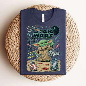 Puede incluir: Una camiseta azul marino con un estampado gr&aacute;fico de Grogu de Star Wars, rodeado de varias naves espaciales, la Estrella de la Muerte, BB-8 y R2-D2 sobre una superficie lunar. El texto Star Wars aparece en una fuente negrita de color verde oscuro.