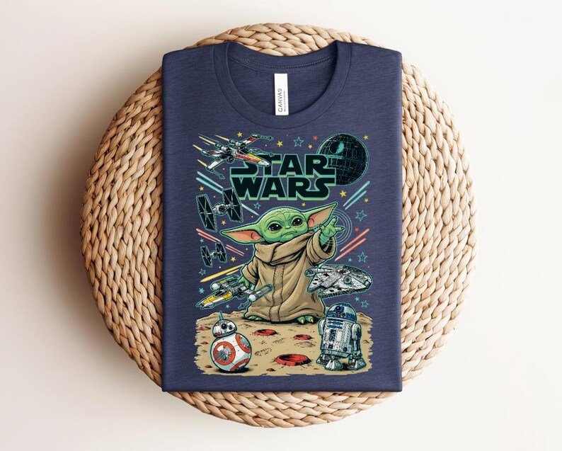 Puede incluir: Una camiseta azul marino con un estampado gr&aacute;fico de Grogu de Star Wars, rodeado de varias naves espaciales, la Estrella de la Muerte, BB-8 y R2-D2 sobre una superficie lunar. El texto Star Wars aparece en una fuente negrita de color verde oscuro.