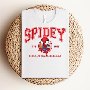 May include: White t-shirt with a red graphic that says "SPIDEY" in a collegiate font. Below the word "SPIDEY" is a cartoon image of Spider-Man and the text "EST 2021" and "SPIDEY AND HIS AMAZING FRIENDS". The shirt is folded on a woven, circular surface.