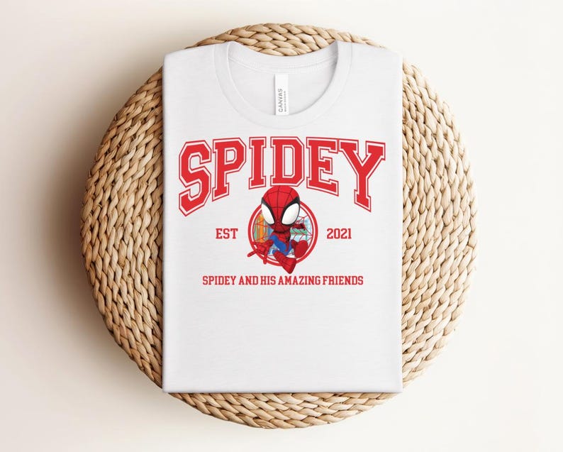 May include: White t-shirt with a red graphic that says "SPIDEY" in a collegiate font. Below the word "SPIDEY" is a cartoon image of Spider-Man and the text "EST 2021" and "SPIDEY AND HIS AMAZING FRIENDS". The shirt is folded on a woven, circular surface.
