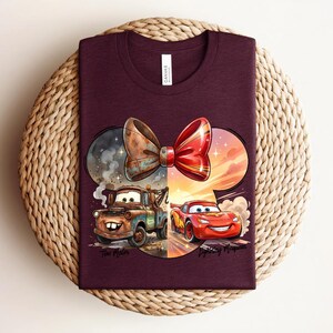 May include: A maroon t-shirt featuring a graphic of a large bow shape containing two cartoon cars. On the left, a rusty brown tow truck labelled 'Tow Mater' is surrounded by smoke. On the right, a red race car labelled 'Lightning McQueen' is driving.