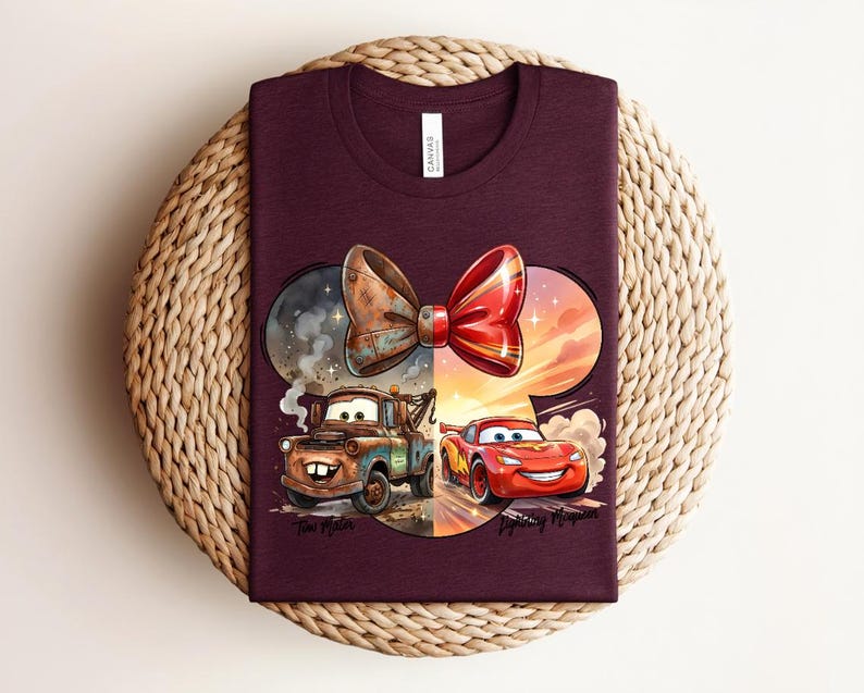 May include: A maroon t-shirt featuring a graphic of a large bow shape containing two cartoon cars. On the left, a rusty brown tow truck labelled 'Tow Mater' is surrounded by smoke. On the right, a red race car labelled 'Lightning McQueen' is driving.