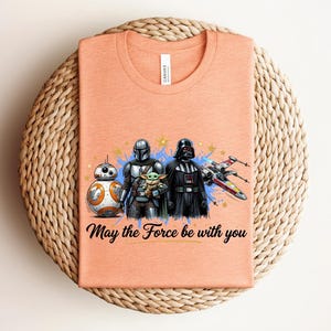 Pode incluir: Uma t-shirt cor de p&ecirc;ssego com uma impress&atilde;o gr&aacute;fica de BB-8, o Mandaloriano, Grogu e Darth Vader ao lado de um ca&ccedil;a estelar X-wing. O texto "May the Force be with you" est&aacute; impresso em letra cursiva preta abaixo das personagens.