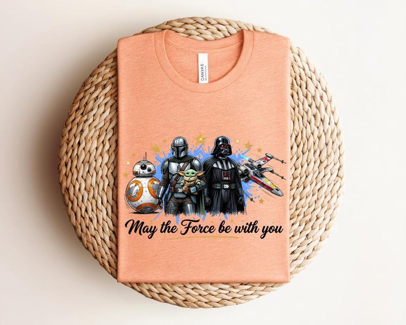 Pode incluir: Uma t-shirt cor de p&ecirc;ssego com uma impress&atilde;o gr&aacute;fica de BB-8, o Mandaloriano, Grogu e Darth Vader ao lado de um ca&ccedil;a estelar X-wing. O texto "May the Force be with you" est&aacute; impresso em letra cursiva preta abaixo das personagens.