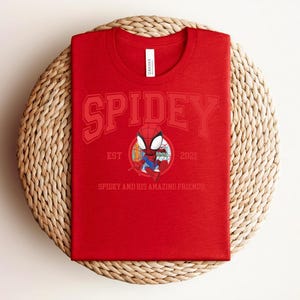 May include: Red t-shirt featuring the word "SPIDEY" in a collegiate font, with an illustration of Spider-Man in a circular emblem. The text "EST 2021" and "SPIDEY AND HIS AMAZING FRIENDS" are also visible. The shirt is displayed on a woven, circular mat.