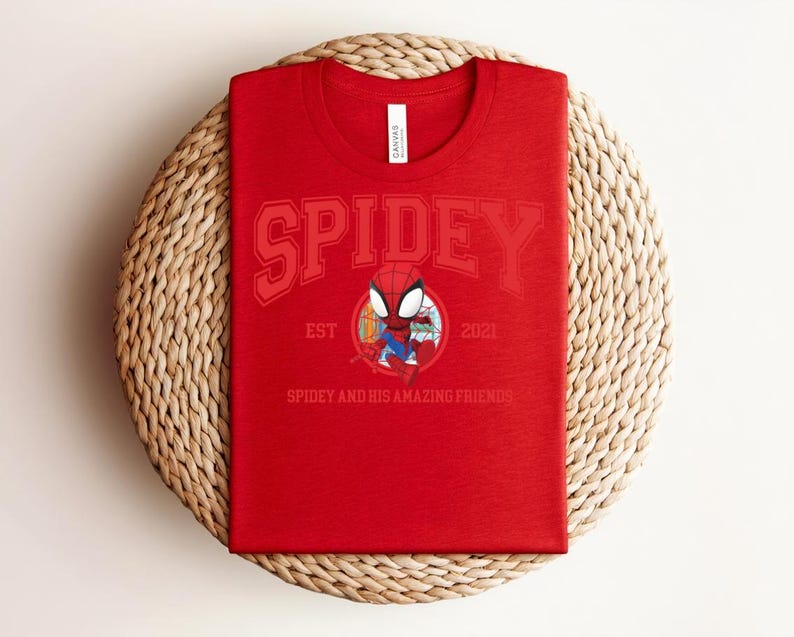 May include: Red t-shirt featuring the word "SPIDEY" in a collegiate font, with an illustration of Spider-Man in a circular emblem. The text "EST 2021" and "SPIDEY AND HIS AMAZING FRIENDS" are also visible. The shirt is displayed on a woven, circular mat.