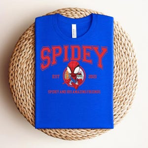 May include: A royal blue t-shirt with the word "SPIDEY" in red arched lettering. Below the word "SPIDEY" is a cartoon image of Spider-Man. The text "EST 2021" and "SPIDEY AND HIS AMAZING FRIENDS" are also printed on the shirt.