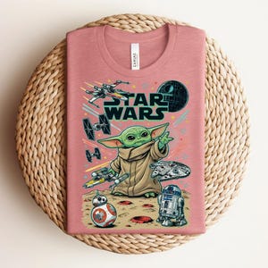 Puede incluir: Una camiseta de cuello redondo en color rosa empolvado con un estampado gr&aacute;fico de Star Wars. El dise&ntilde;o incluye a Grogu, BB-8, R2-D2, el Halc&oacute;n Milenario, un X-wing, cazas TIE y la Estrella de la Muerte. El texto STAR WARS aparece en may&uacute;sculas.