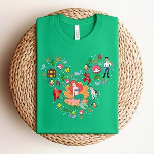 K&ouml;nnte beinhalten: Ein gr&uuml;nes T-Shirt mit einem herzf&ouml;rmigen Design, das eine Meerjungfrau, einen Prinzen und andere Charaktere zeigt. Die Grafik enth&auml;lt Meereskreaturen und florale Elemente. Das T-Shirt liegt gefaltet auf einer gewebten Oberfl&auml;che.