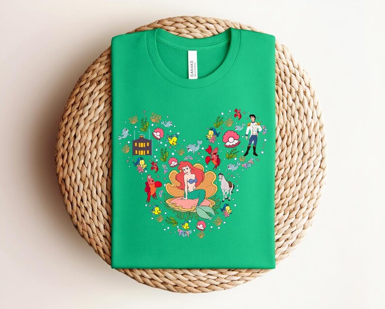 K&ouml;nnte beinhalten: Ein gr&uuml;nes T-Shirt mit einem herzf&ouml;rmigen Design, das eine Meerjungfrau, einen Prinzen und andere Charaktere zeigt. Die Grafik enth&auml;lt Meereskreaturen und florale Elemente. Das T-Shirt liegt gefaltet auf einer gewebten Oberfl&auml;che.