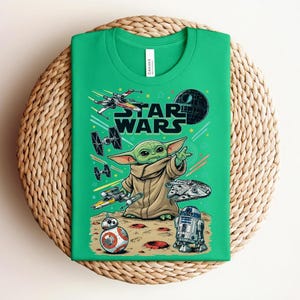 Puede incluir: Una camiseta de color verde brillante con un estampado gr&aacute;fico de Star Wars. El dise&ntilde;o incluye a Grogu, la Estrella de la Muerte, un X-wing, cazas TIE, el Halc&oacute;n Milenario, BB-8 y R2-D2 sobre una superficie arenosa con l&iacute;neas de movimiento y estrellas.