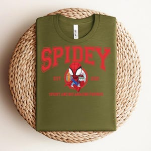 May include: Olive green t-shirt featuring a red graphic of Spider-Man with the word "SPIDEY" above and "SPIDEY AND HIS AMAZING FRIENDS" below. The shirt is displayed on a woven, circular placemat.