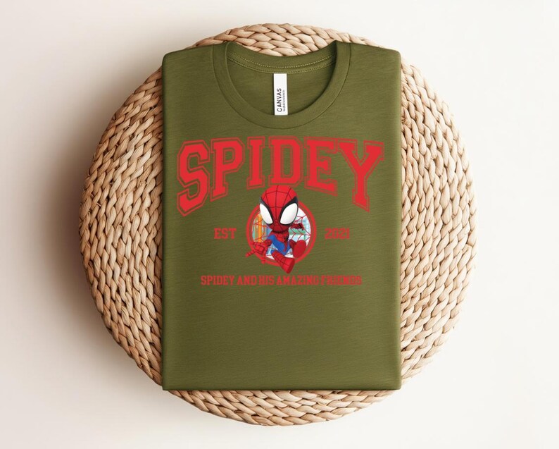May include: Olive green t-shirt featuring a red graphic of Spider-Man with the word "SPIDEY" above and "SPIDEY AND HIS AMAZING FRIENDS" below. The shirt is displayed on a woven, circular placemat.