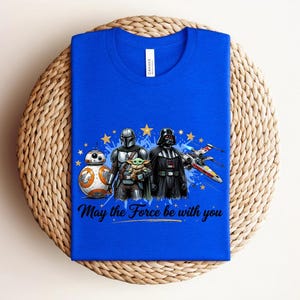 Pode incluir: Uma t-shirt azul royal com uma estampa gr&aacute;fica de BB-8, O Mandaloriano, Grogu e Darth Vader ao lado de um ca&ccedil;a estelar X-wing. O design inclui estrelas amarelas, salpicos de tinta azul e o texto "May the Force be with you" em preto.