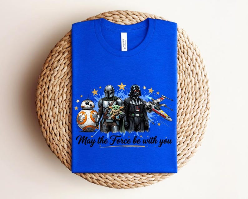 Pode incluir: Uma t-shirt azul royal com uma estampa gr&aacute;fica de BB-8, O Mandaloriano, Grogu e Darth Vader ao lado de um ca&ccedil;a estelar X-wing. O design inclui estrelas amarelas, salpicos de tinta azul e o texto "May the Force be with you" em preto.