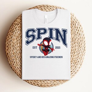 K&ouml;nnte beinhalten: Wei&szlig;es T-Shirt mit einem marineblauen Grafikdesign. Das Wort "SPIN" ist in einer College-Schriftart gehalten. Darunter befindet sich ein Cartoon-Superheldenbild mit dem Text "EST 2021" und "SPIDEY AND HIS AMAZING FRIENDS". Das T-Shirt wird auf einer gewebten Matte pr&auml;sentiert.
