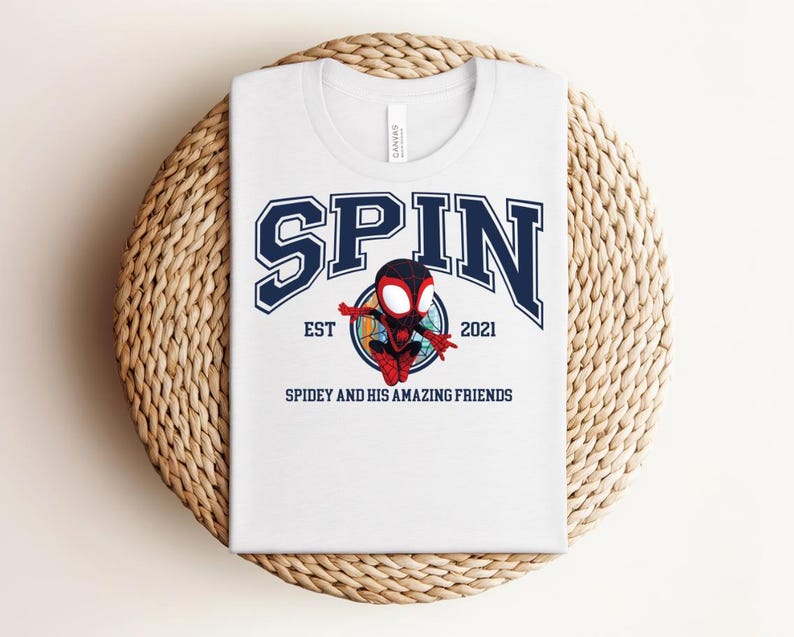 K&ouml;nnte beinhalten: Wei&szlig;es T-Shirt mit einem marineblauen Grafikdesign. Das Wort "SPIN" ist in einer College-Schriftart gehalten. Darunter befindet sich ein Cartoon-Superheldenbild mit dem Text "EST 2021" und "SPIDEY AND HIS AMAZING FRIENDS". Das T-Shirt wird auf einer gewebten Matte pr&auml;sentiert.