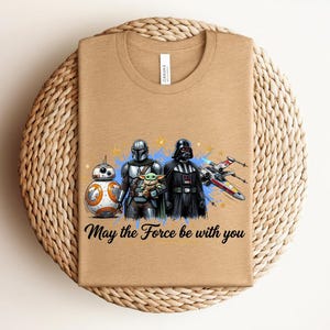 Pode incluir: Uma t-shirt de gola redonda bege com uma estampa gr&aacute;fica de BB-8, o Mandaloriano, Grogu e Darth Vader ao lado de um ca&ccedil;a estelar X-wing. O texto "May the Force be with you" est&aacute; impresso em preto sob a ilustra&ccedil;&atilde;o colorida das personagens.
