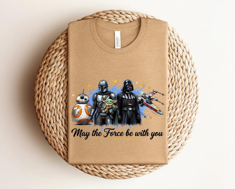 Pode incluir: Uma t-shirt de gola redonda bege com uma estampa gr&aacute;fica de BB-8, o Mandaloriano, Grogu e Darth Vader ao lado de um ca&ccedil;a estelar X-wing. O texto "May the Force be with you" est&aacute; impresso em preto sob a ilustra&ccedil;&atilde;o colorida das personagens.