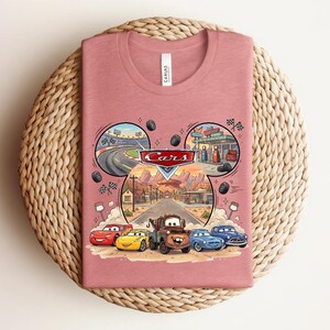 Puede incluir: Una camiseta de color rosa polvoriento con un gr&aacute;fico del logotipo y los personajes de Cars de Disney Pixar, incluyendo a Rayo McQueen y Mate. El dise&ntilde;o incluye un marco con forma de orejas de Mickey con escenas de una pista y Radiator Springs.
