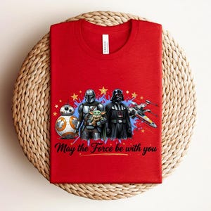 Pode incluir: Uma t-shirt vermelha com uma estampa gr&aacute;fica de BB-8, o Mandaloriano, Grogu e Darth Vader ao lado de um ca&ccedil;a estelar X-wing. O design inclui estrelas amarelas, salpicos de tinta azul e o texto May the Force be with you em preto.