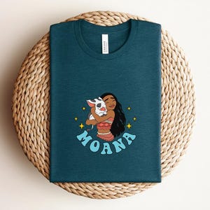 Puede incluir: Una camiseta de cuello redondo en color verde azulado oscuro con un estampado gr&aacute;fico de un personaje de pelo largo y oscuro abrazando a un peque&ntilde;o cerdo manchado. El dise&ntilde;o incluye estrellas amarillas y la palabra MOANA en letras azul claro.