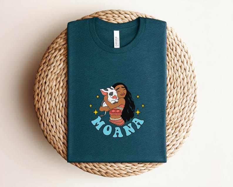 Puede incluir: Una camiseta de cuello redondo en color verde azulado oscuro con un estampado gr&aacute;fico de un personaje de pelo largo y oscuro abrazando a un peque&ntilde;o cerdo manchado. El dise&ntilde;o incluye estrellas amarillas y la palabra MOANA en letras azul claro.
